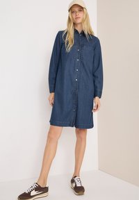 Jeansklänning i mörkblått, långa ärmar, framknäppning, två bröstfickor och rak skärning. Bärs med bruna och vita sneakers.