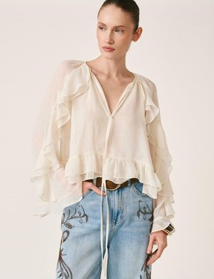 Mujer con blusa transparente color crema con mangas y bajo con volantes, combinada con jeans azul claro bordados y cinturón marrón, con la mano en el bolsillo.