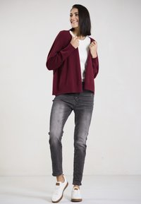 Burgunderfarbener Cardigan mit gerippten Kanten über einem weißen Top, kombiniert mit grauen Jeans und weißen Sneakers mit braunen Akzenten, vor einem neutralen Hintergrund stehend.