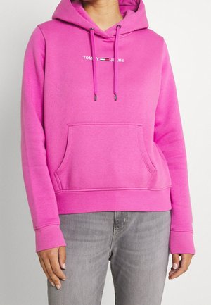 Frau trägt einen leuchtend pinken Hoodie von Tommy Jeans mit Fronttasche und Kordelzug, kombiniert mit hellgrauen Jeans.