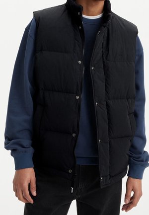 Homme portant une veste matelassée noire par-dessus un sweat-shirt bleu à manches longues et un pantalon noir, debout devant un fond blanc uni.