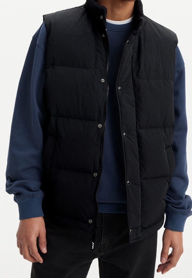 Homme portant une veste matelassée noire par-dessus un sweat-shirt bleu à manches longues et un pantalon noir, debout devant un fond blanc uni.