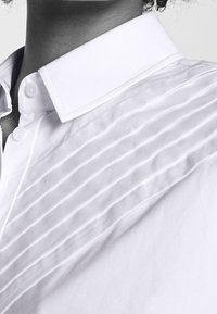 Primo piano di una persona che indossa una camicia bianca con colletto ripiegato e dettagli in tessuto plissettato diagonale sulla spalla.