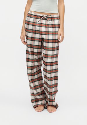 Femme portant un pantalon de pyjama ample en flanelle blanche avec un motif à carreaux rouges et verts et une taille à cordon, debout pieds nus sur fond blanc.