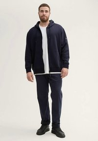 Marineblauwe hoodie met ritssluiting van zachte stof, gecombineerd met een wit T-shirt en donkermarineblauwe broek. Zwarte sneakers maken de outfit compleet. Eenvoudig ontwerp.