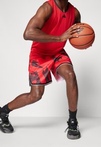 Débardeur de sport rouge, short rouge avec motif triangulaire noir, baskets noires et grises. Tenant un ballon de basketball orange.