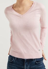 Woman wearing a light pink V-neck sweater and dark blue jeans, with a small white Polo Club label near the hem on the right side.