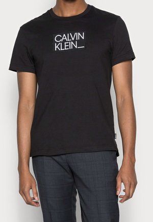 Mężczyzna ubrany w czarną koszulkę z krótkim rękawem Calvin Klein z białym logo na środku klatki piersiowej i ciemne spodnie w kratę.