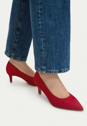 Barfuß mit leuchtend roten Wildleder-Pumps mit spitzer Zehenpartie und niedrigem Absatz, kombiniert mit blauen, verkürzten Jeans vor weißem Hintergrund.