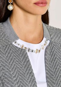 Vrouw in pareldruppel oorbellen, juweel-witte top en grijs jaquard jasje dat de nek en lippen toont.