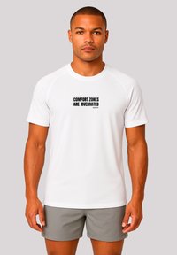 White short-sleeve athletic shirt with black text reading "COMFORT ZONES ARE OVERRATED" across the chest. Smooth fabric, round neck.