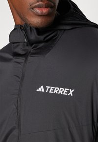 Crna lagana jakna s ovratnikom na patent zatvarač, s bijelim "TERREX" logom na prednjoj strani. Materijal izgleda glatko i izdržljivo.