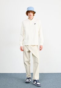 Polo de manga longa em creme com um logótipo preto da Nike, combinado com calças off-white e um chapéu de balde azul, apresentando um design texturizado.