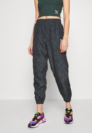 Frau trägt dunkel gemusterte Puma-Jogginghosen mit Blumenmuster, schwarzen Crop-Top und farbenfrohe schwarze, pinke, orange und violette Turnschuhe, steht vor einfachem Hintergrund.