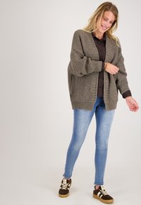 Chunky, gestructureerde olijfgroene cardigan met geribde manchetten en zoom, gedragen over een donker, knoopsluitend shirt en lichte skinny jeans. Bruine sneakers.