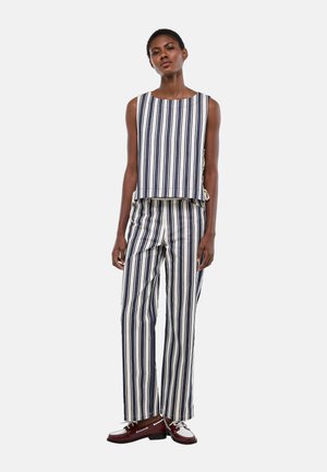 Femme portant un haut sans manches blanc et un pantalon assorti avec des rayures verticales bleu marine, assortis à des mocassins blancs et bordeaux, debout.