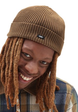 Gorro de punto marrón con textura acanalada, con una etiqueta de logo negra de Vans. El sujeto tiene dreadlocks largos y estilizados y lleva una camisa de cuadros.