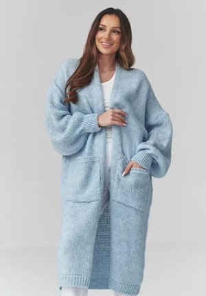 Kobieta ubrana w długi, jasnoniebieski, gruby, dziergany kardigan z dużymi kieszeniami, na białej bluzce i spodniach, uśmiechająca się z ręką w kieszeni.