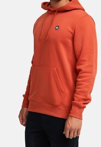 Orange hoodie gjord av mjuk tyg, med en framfickan för händerna, justerbar huva med dragsko och en liten logotyp på bröstet.