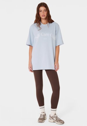 Młoda kobieta ubrana w jasnoniebieski, szeroki t-shirt z napisem "Self Care Club", brązowe legginsy, białe skarpetki i beżowe sneakersy, stojąca na białym tle.