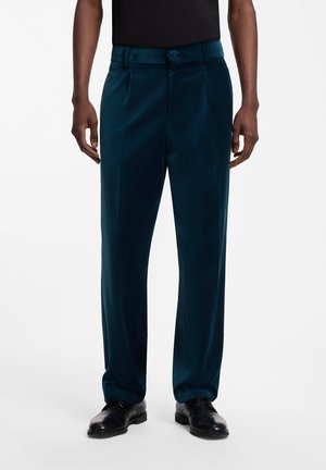 Mannelijk model draagt turquoise fluwelen pantalon en zwarte nette schoenen, staand tegen een effen witte achtergrond.