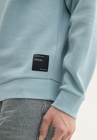 Felpa azzurra chiaro con polsini e orlo a coste, con un'etichetta nera che riporta il nome del marchio e informazioni sulla qualità.