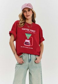 Czerwona bawełniana koszulka z podwiniętymi rękawami, z grafiką ekspresu do kawy i napisem. Połączona z jasnoniebieskimi spodniami o szerokich nogawkach. Różowa czapka.