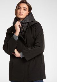 Zwarte geïsoleerde jas met een afneembare glanzende capuchon. Heeft een voetzak, zijzakken en een zachte binnenvoering.