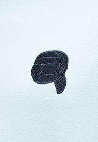 Μαύρο κεντημένο λογότυπο με πρόσωπο που φοράει κράνος και προσωπίδα σε ανοιχτό μπλε ύφασμα.