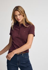 Burgundowa koszulka polo z krótkim rękawem, z zapięciem na trzy guziki i haftowanym logo na piersi, w połączeniu z ciemnoniebieskimi dżinsami i brązowym paskiem.