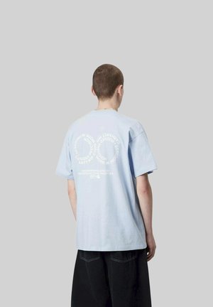 Persona con capelli corti che indossa una maglietta oversize azzurro chiaro con testo bianco circolare sul retro, abbinata a pantaloni neri larghi, sfondo grigio.