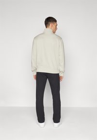 Ljus beige zip-up sweatshirt med hög krage, ribbade muddar och fåll, kombinerad med mörkgrå byxor och vita sneakers, sedd bakifrån.