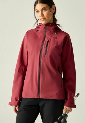 Vrouw die een rode capuchonjas en zwarte broek draagt, een wandelstok vasthoudt en iets opzij kijkt tegen een egale achtergrond.