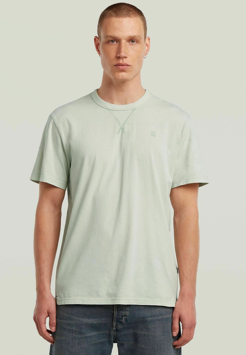 T-shirt di cotone verde chiaro con collo a giro, maniche corte e dettagli di cucitura incrociata sottili sul collo, abbinata a jeans blu scuro.