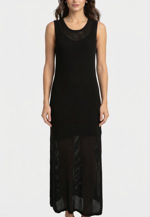 Robe maxi noire sans manches en maille avec une partie inférieure transparente, portée par une femme debout aux cheveux lâchés et aux bijoux minimalistes.