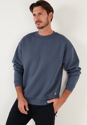 Blå bomulds sweatshirt med rund hals, faldne skuldre og ribbede manchetter. Har et lille mærke på den nederste side. Blød tekstur.