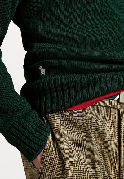 Groen gebreid trui met ribbelzoom en manchet, voorzien van een klein geborduurd paardenlogo. Gecombineerd met bruine geruite broek.