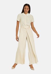 Crème-kleurige tweedelige outfit met een blouse met korte mouwen en kraag, en een broek met brede pijpen en een gedraaide voorkant. Soepele textuur.