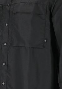 Zwarte overhemdd blouse met een zak en een gestructureerde stof. Duurzaam materiaal met drukknoopsluiting en een klein logo-label aan de zijkant.