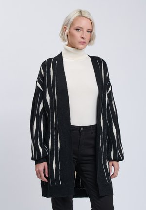 Frau mit kurzen blonden Haaren, die einen weißen Rollkragenpullover, eine schwarz-weiß gestreifte Strickjacke und schwarze Hose vor einem einfarbigen Hintergrund trägt.
