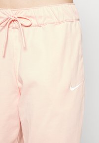 Gaiši rozā sporta bikses ar elastīgu jostu, regulējamu auklu un mazu balto Nike logo kreisajā augšstilbā. Mīksta auduma tekstūra.