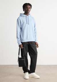 Sudadera azul clara con cordones, combinada con pantalones de mezclilla oscuros y zapatillas blancas. El modelo sostiene un pequeño bolso negro cruzado.