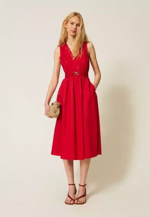 Donna con capelli biondi lunghi che indossa un vestito midi rosso senza maniche e sandali rossi, tenendo in una mano una piccola pochette intrecciata beige.