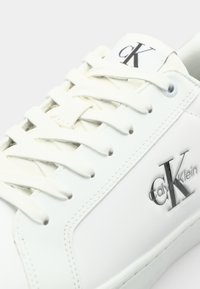 Baskets en cuir blanc avec des lacets blancs, arborant un logo "cK" argenté et une texture lisse. Le panneau latéral présente un détail de couture subtil.
