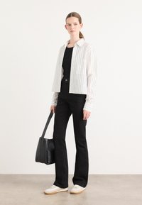 Veste courte à rayures blanches portée sur un haut noir, associée à un jean noir évasé et des baskets beiges. Un sac à main noir est tenu d'une main.
