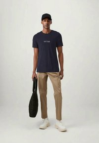 Mörkblå bomullströja med en vit "GU3SS" logotyp, beige byxor, vita sneakers och en svart vadderad väska. Modellen står mot en ljus bakgrund.