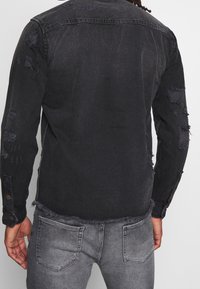 Chaqueta de mezclilla negra con bordes deshilachados y detalles desgastados. Mangas largas con puños abotonados. Combinada con jeans grises.