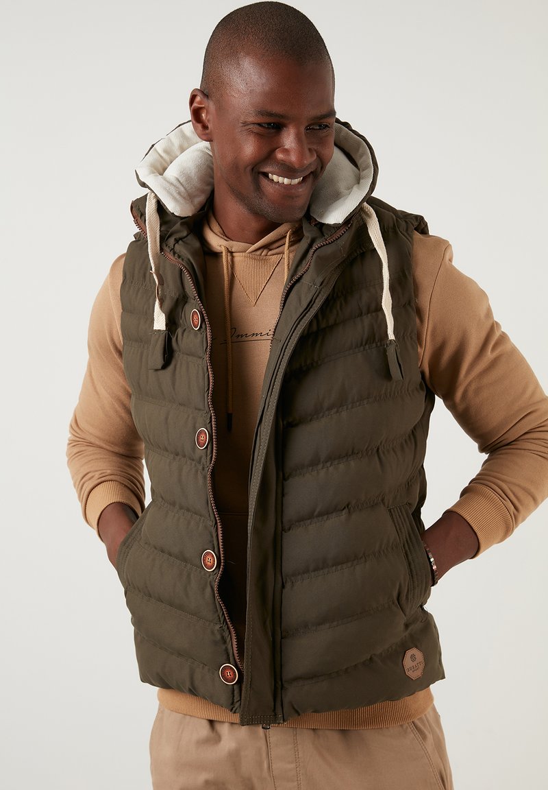 Padded olive green vest with a hood, featuring a zipper and brown buttons, worn over a tan sweatshirt. Notable quilted texture and logo patch.