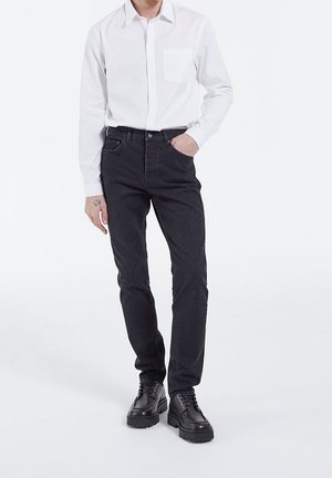 Persona che indossa jeans neri aderenti, scarpe nere con lacci e una camicia bianca a maniche lunghe con bottoni, in piedi con una mano in tasca.