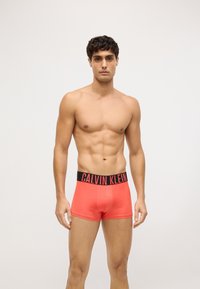 Crvene Calvin Klein bokserice s crnim rubom u struku, koje imaju prilagodljiv kroj i glatku teksturu. Nema dodatne odjeće ili dodataka vidljivih.